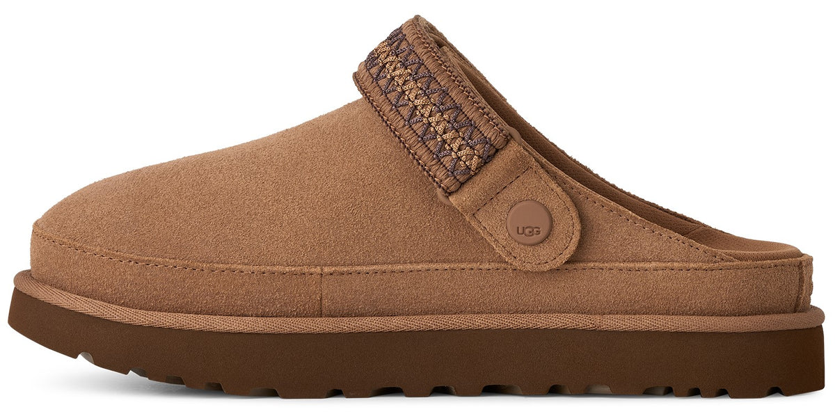 A tan suede slip-on shoe with a thick, dark brown textured platform sole and a strap across the top featuring a decorative, patterned woven band and a small round tab embossed with the UGG logo.