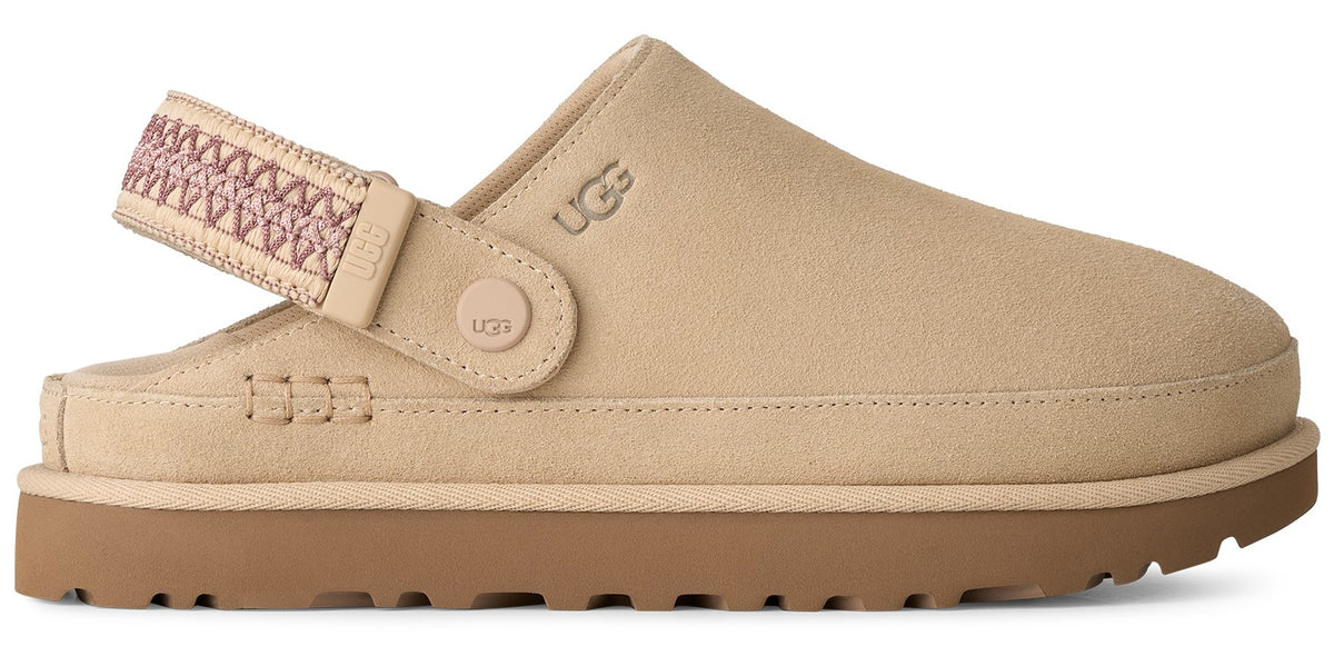 A tan suede slip-on shoe with a round toe and a brown rubber platform sole, featuring an adjustable beige woven heel strap with a small tan circular logo button and the word UGG embossed on the side.