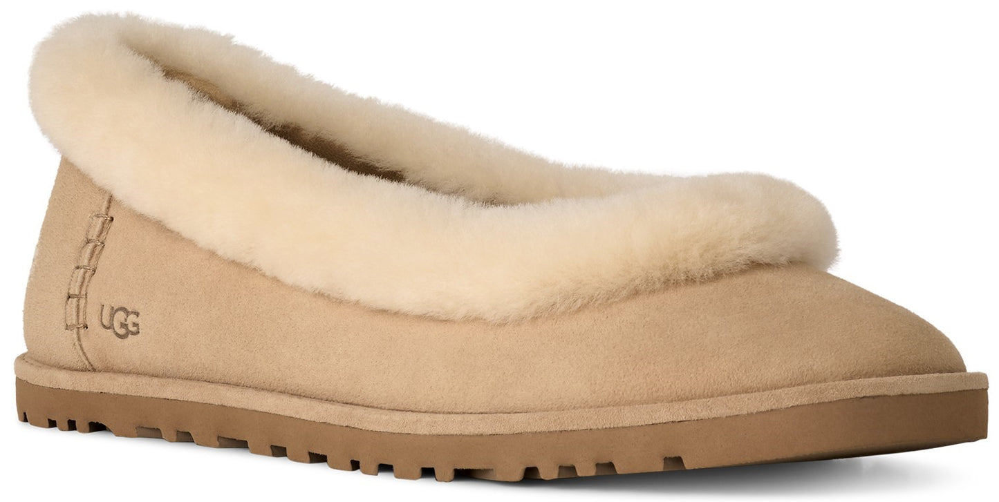 A tan suede slip-on shoe with a thick, fluffy cream-colored sheepskin collar and a brown rubber sole, featuring the brand name UGG embossed on the heel area.