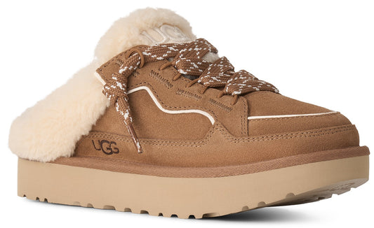 A tan suede platform slip-on shoe with a cream-colored shearling lining at the heel, featuring patterned laces and the text UGG embossed on the side.