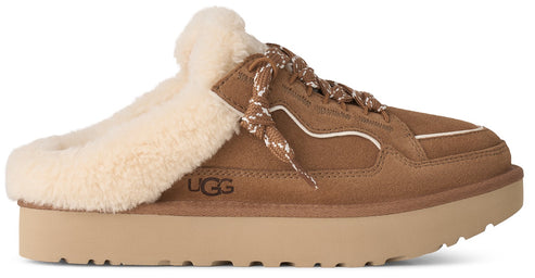 A brown suede slip-on shoe with a thick tan platform sole and a fluffy off-white shearling collar, featuring decorative laces and the word UGG embossed on the side.