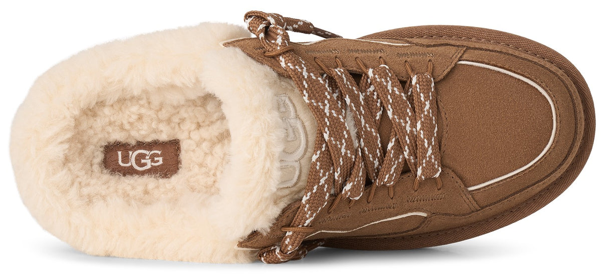 A tan suede slip-on shoe with a cream-colored shearling lining, featuring brown and white patterned laces, and the text UGG on the interior footbed.