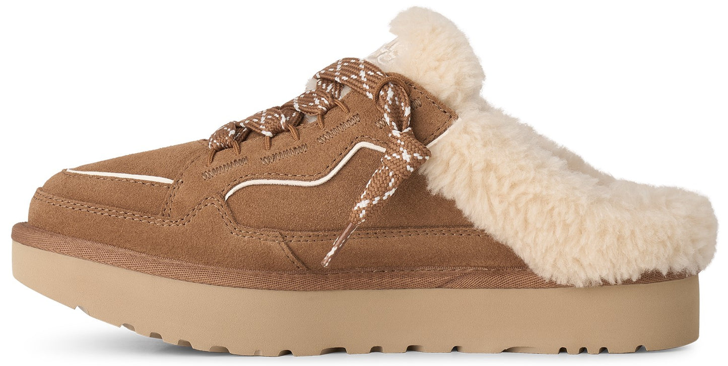A tan suede platform slip-on mule with a thick rubber sole, featuring a cream-colored shearling collar, white decorative piping, and a patterned lace-up front detail.