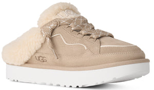 A tan suede slip-on shoe with a thick white platform sole and cream-colored faux fur lining. It features decorative laces and the word UGG debossed on the side and tongue.