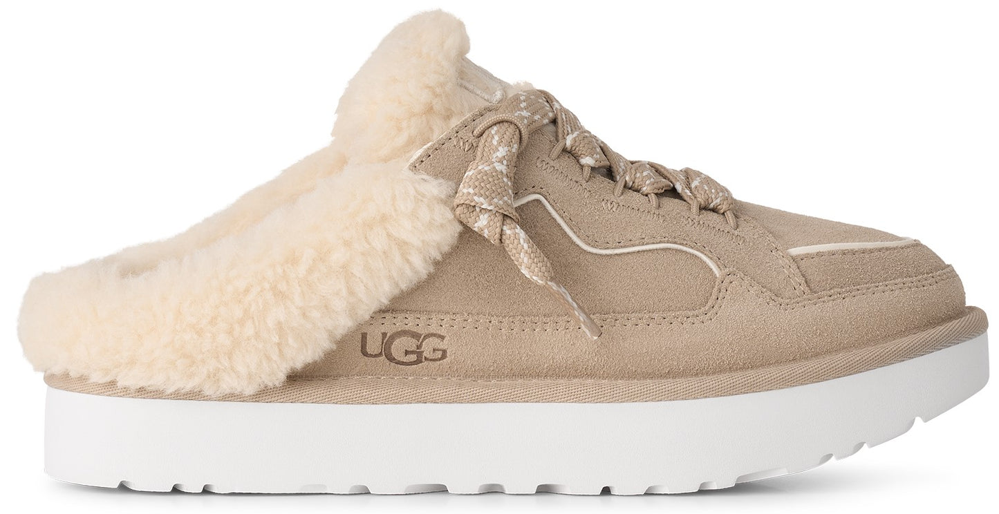 A tan suede slip-on shoe with a thick white platform sole and a fluffy cream-colored shearling lining, featuring decorative laces on the top and the text UGG embossed on the side.