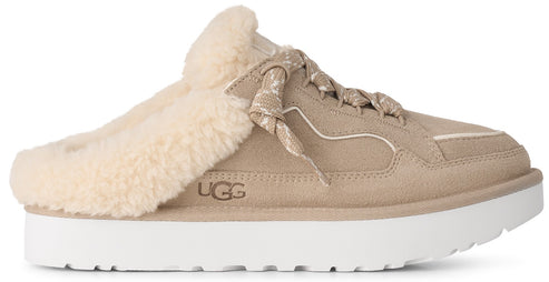 A tan suede slip-on shoe with a thick white platform sole and a fluffy cream-colored shearling lining, featuring decorative laces on the top and the text UGG embossed on the side.