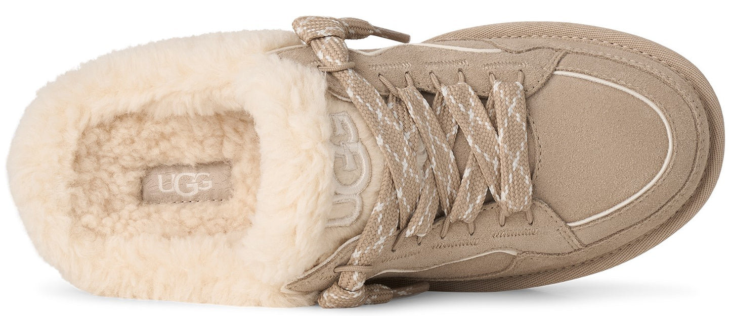 A tan slip-on shoe with a cream-colored shearling lining, featuring decorative crisscrossed laces on top and the brand name embossed on the insole.