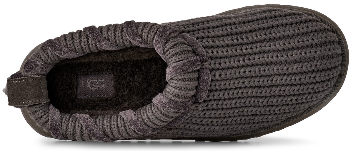 A top-down view of a dark grey, ribbed knit slip-on slipper with a plush, fuzzy interior and a visible UGG logo patch on the footbed.