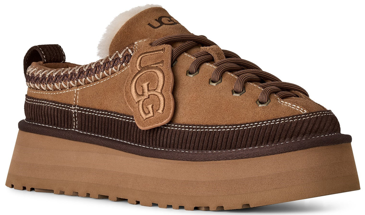 A tan suede platform sneaker with dark brown ribbed detailing around the midsole, dark brown laces, an UGG branded tag, a white shearling-lined collar, and a thick, chunky tan sole.