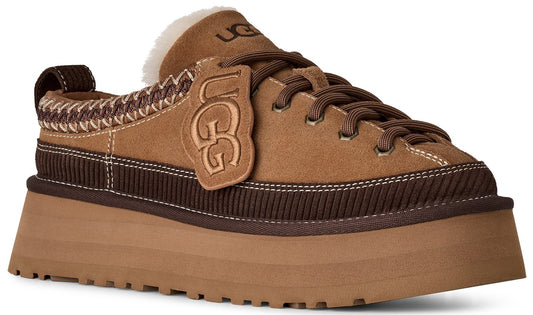A tan suede platform sneaker with dark brown ribbed detailing around the midsole, dark brown laces, an UGG branded tag, a white shearling-lined collar, and a thick, chunky tan sole.