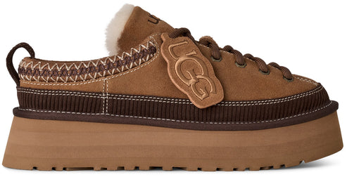 A brown suede platform slip-on shoe with a lace-up front, a brown ribbed midsole, a thick tan rubber sole, and a decorative leather tag with the embossed letters UGG attached to the side.