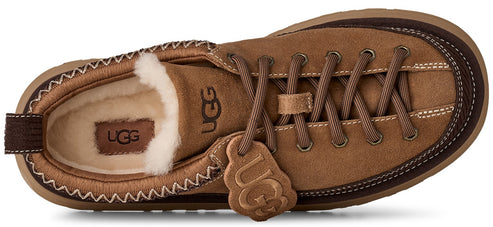 A top-down view of a tan suede low-top shoe with cream-colored faux fur lining, brown laces, a brown rubber sole, and a UGG logo patch on the side and heel bed.