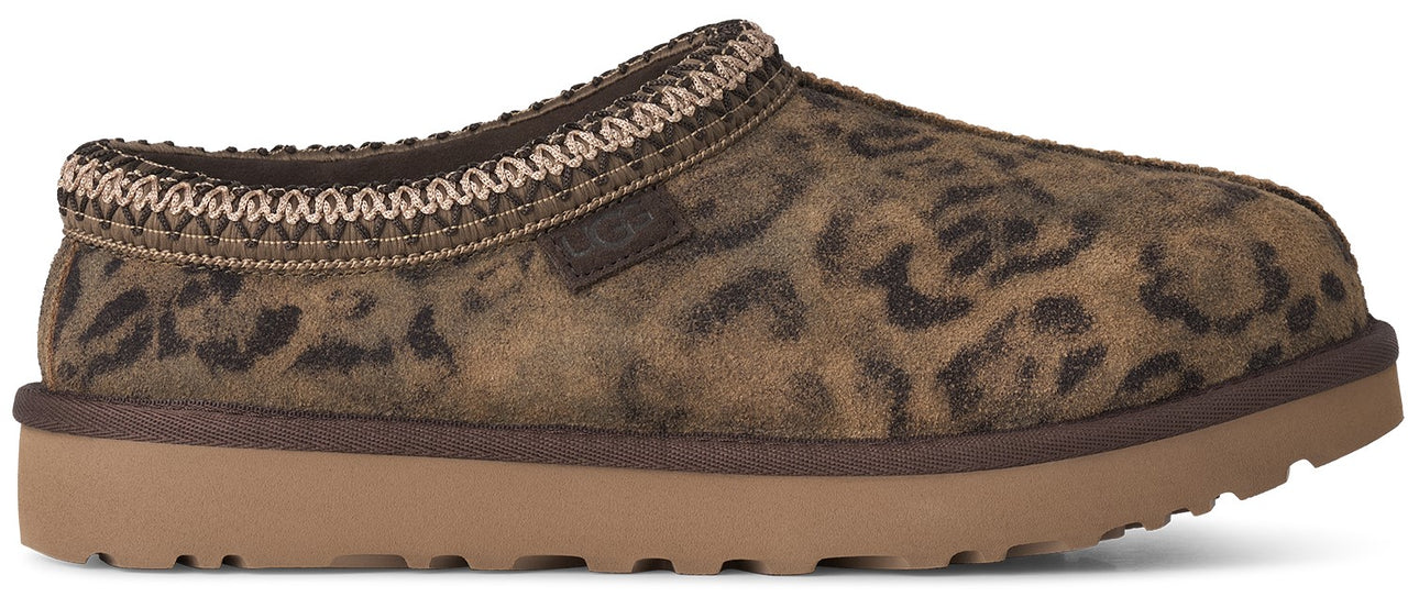 A slip-on shoe with a brown leopard print suede upper and a thick, flat, light brown rubber outsole, featuring a decorative braided stitch trim around the collar and a small label embossed with "UGG" on the side.