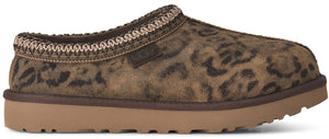 A slip-on shoe with a brown leopard print suede upper and a thick, flat, light brown rubber outsole, featuring a decorative braided stitch trim around the collar and a small label embossed with "UGG" on the side.