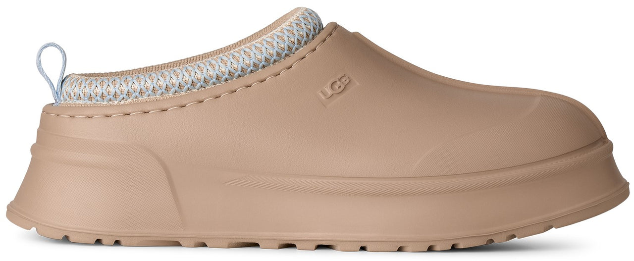 A tan slip-on shoe with a thick platform sole, a rounded toe, a light blue knitted collar, and a pull tab at the heel, featuring the brand name UGG embossed on the side.