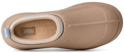 A top-down view of a light tan slip-on shoe with a rounded toe, featuring a plush, light-colored fleece-lined interior collar with a patterned trim and a small rectangular tab displaying the word UGG in the heel area.