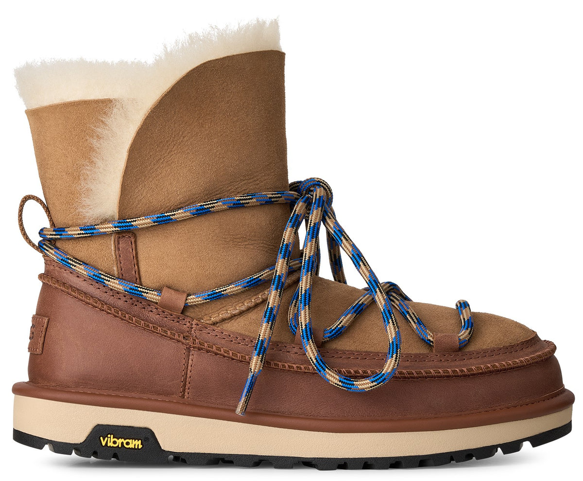 A brown leather and suede winter boot featuring a white shearling cuff, blue and tan rope laces that wrap around the ankle, a thick cream-colored midsole, and a black Vibram outsole.