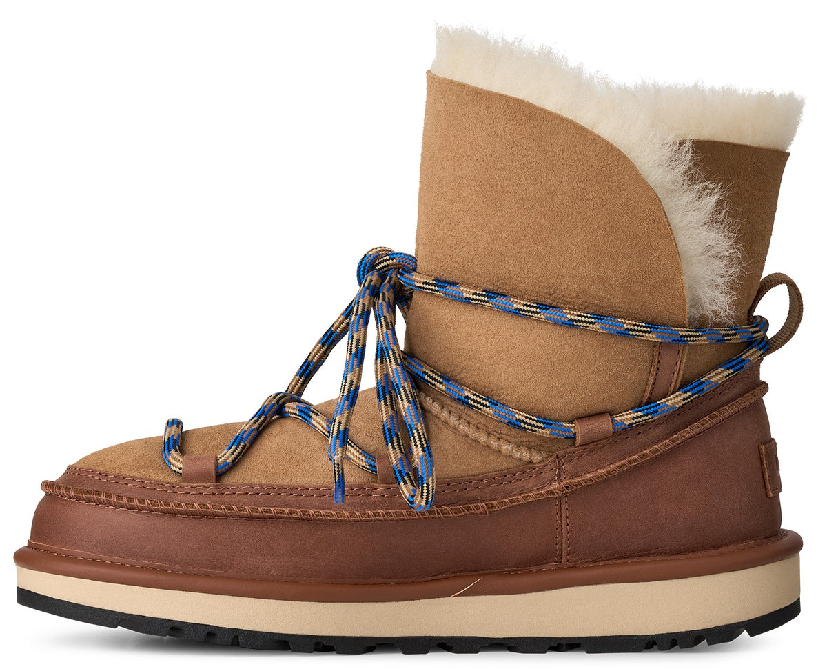 A tan suede winter boot with a brown leather lower trim, thick cream-colored sole, fluffy white shearling lining, and crisscrossing blue and tan patterned laces wrapped around the ankle.