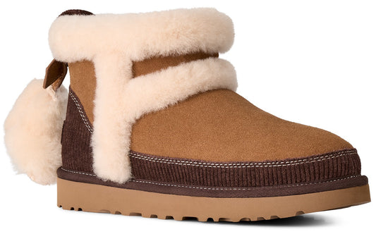 A tan suede ankle boot featuring thick, cream-colored shearling lining around the collar and a decorative trim, a dark brown ribbed texture detail around the base, a thick rubber sole, and a fluffy pom-pom attached to the heel area.