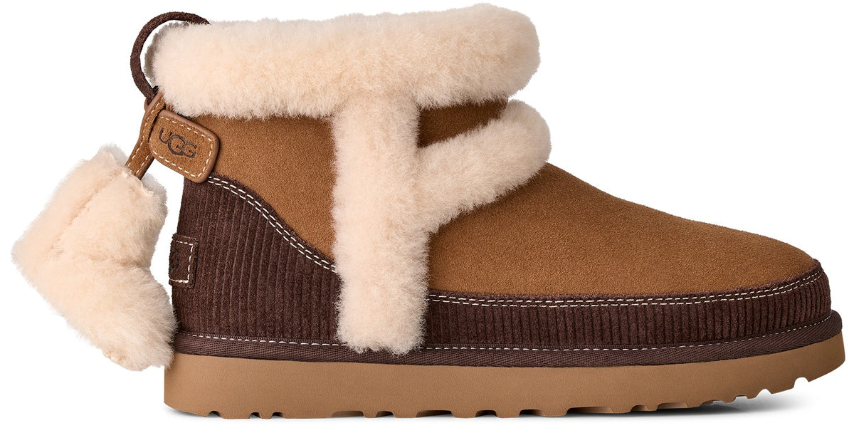 A tan and dark brown suede ankle boot with thick cream-colored sheepskin lining around the collar and a vertical accent stripe, featuring a dark brown ribbed panel on the heel and a fuzzy decorative charm hanging from a heel pull tab with the embossed text UGG.