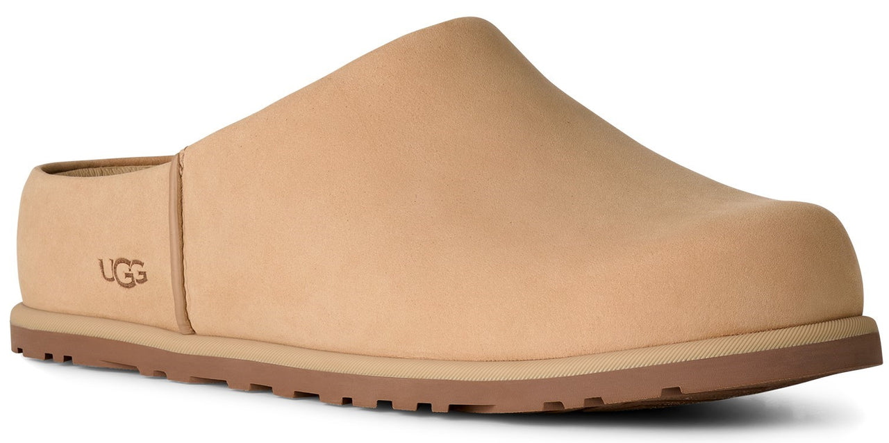 A tan-colored slip-on clog with a smooth leather-like finish and a thick brown treaded sole, featuring the word UGG embossed on the side near the heel.