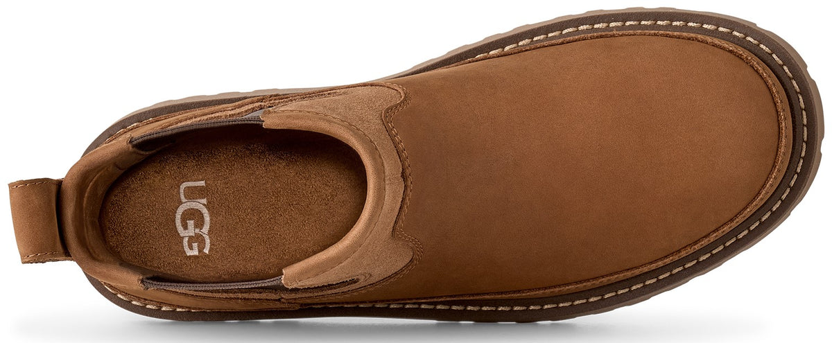 A top-down view of a tan suede slip-on shoe with white contrast stitching along the sole edge and the text UGG visible on the insole.