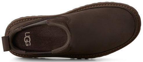 A top-down view of a dark brown suede slip-on shoe with a rounded toe, featuring the UGG logo printed on the interior footbed.