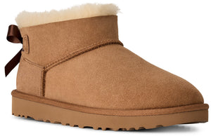 A tan suede ankle boot with a beige shearling lining at the cuff and a small brown ribbon bow attached at the back heel.