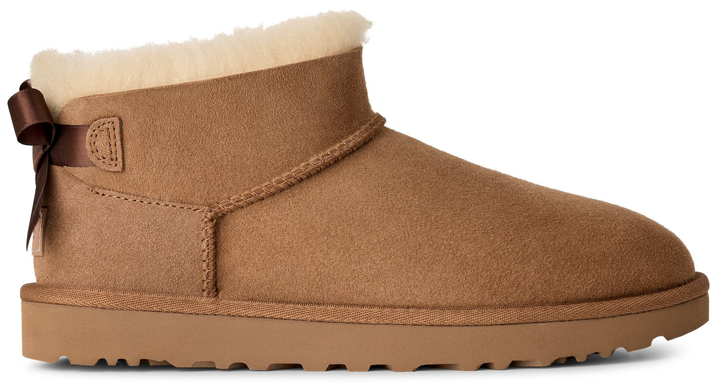 A tan suede ankle boot with a cream-colored shearling lining, featuring a small brown decorative ribbon bow attached to the heel area and a matching flat tan rubber sole.
