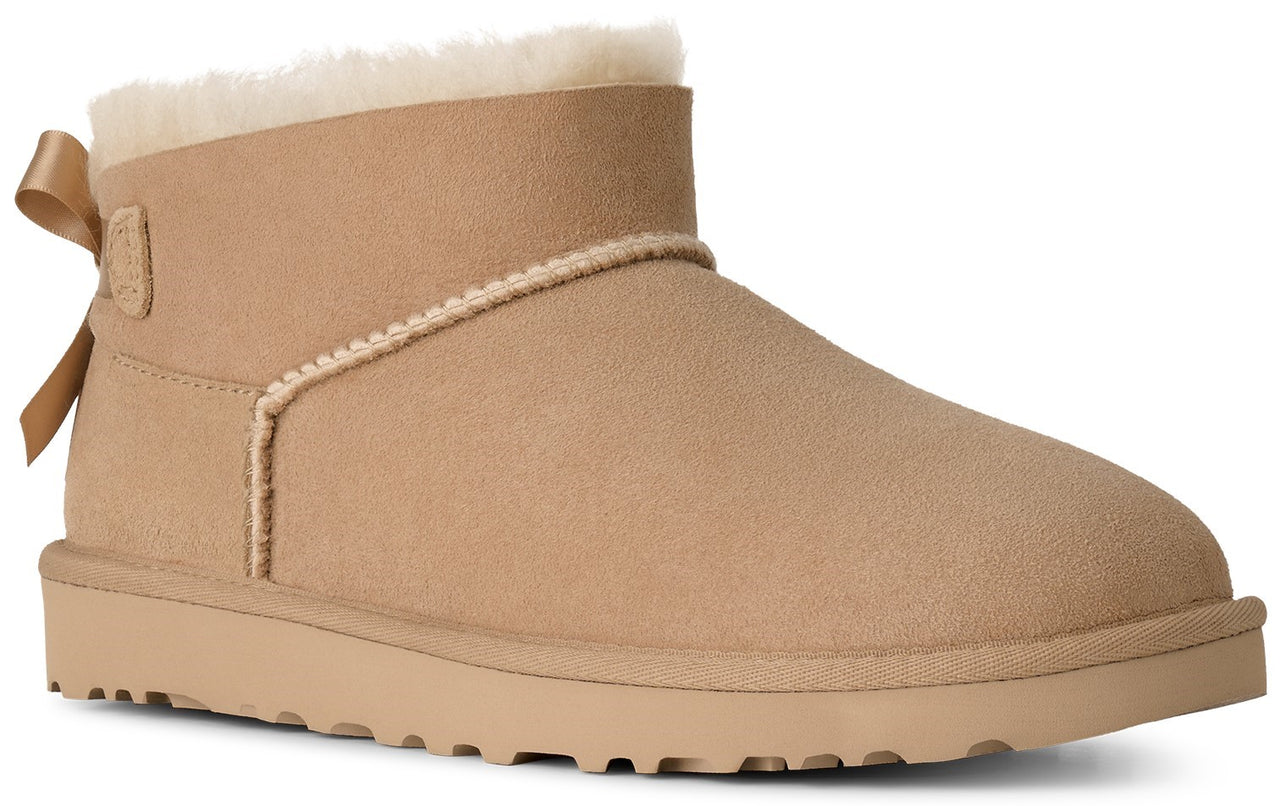 A tan suede ankle boot featuring a fluffy shearling collar and a matching tan ribbon bow at the heel.