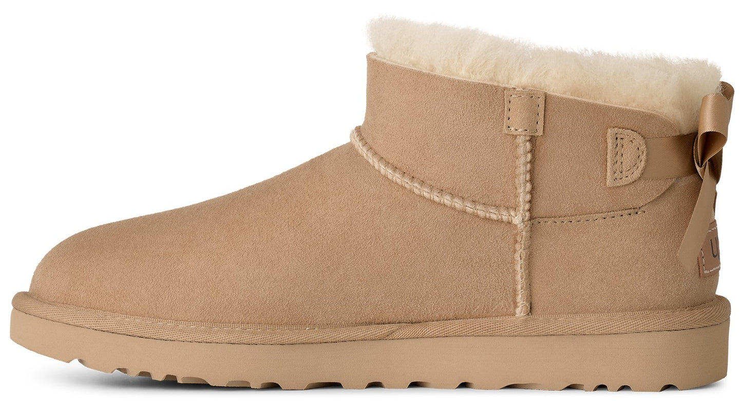 A tan suede ankle boot featuring a thick beige faux-fur lining at the cuff and a small decorative bow detail on the back heel.