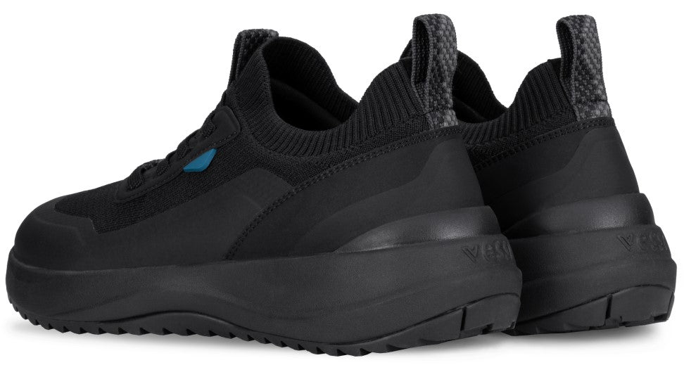 A pair of black Vessi slip-on sneakers seen from a rear three-quarter angle, featuring a textured knit upper, thick matching black soles, and patterned pull tabs on the heels.