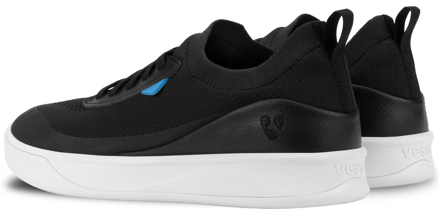 A pair of black knit sneakers with a thick white sole, showing a partial side and rear view, with a small blue accent tag near the laces and a logo debossed on the outer heel area.