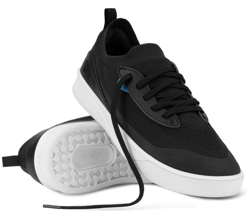 A pair of black low-top sneakers with white soles, one shown from the side and the other showing the white tread pattern on the bottom, featuring black laces and subtle blue accents on the eyelets.