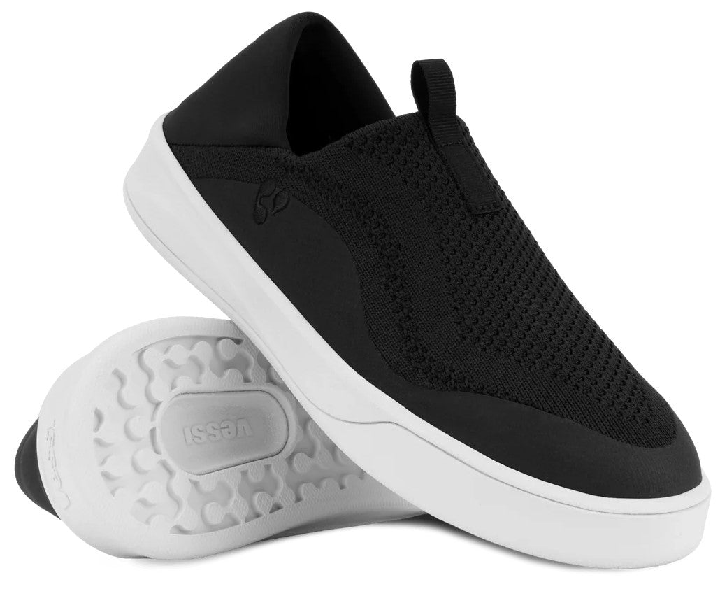 A black slip-on sneaker with a white rubber sole. The shoe features a textured, mesh-like upper and a pull tab on the heel. Another shoe is positioned underneath, showing its white, textured tread pattern with the word 'vessi' embossed in the center.