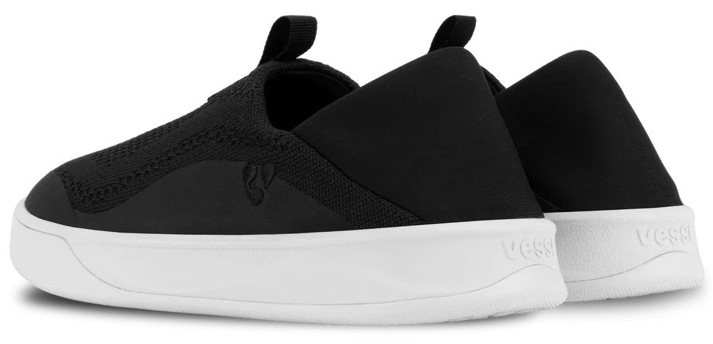 A pair of black slip-on sneakers with a thick white rubber sole, featuring a pull tab on the heel and the text Vessi debossed on the side of the heel.