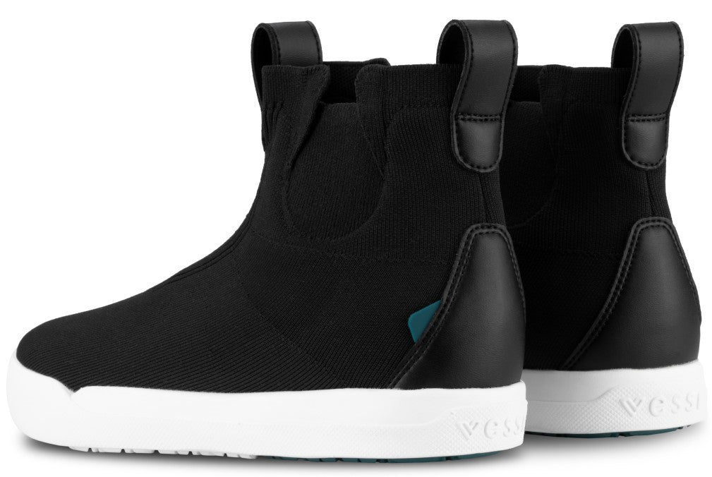 A pair of black slip-on ankle boots with thick white soles and pull tabs on the back, featuring a small teal accent near the heel and the word Vessi embossed on the side of the heel.