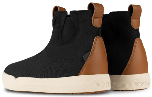 A pair of black ankle boots with tan leather-colored accents on the heel and collar, featuring a thick off-white rubber sole with the word Vessi embossed on the side.