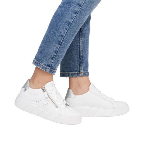 A person wearing blue jeans and white sneakers with silver accents and side zippers.