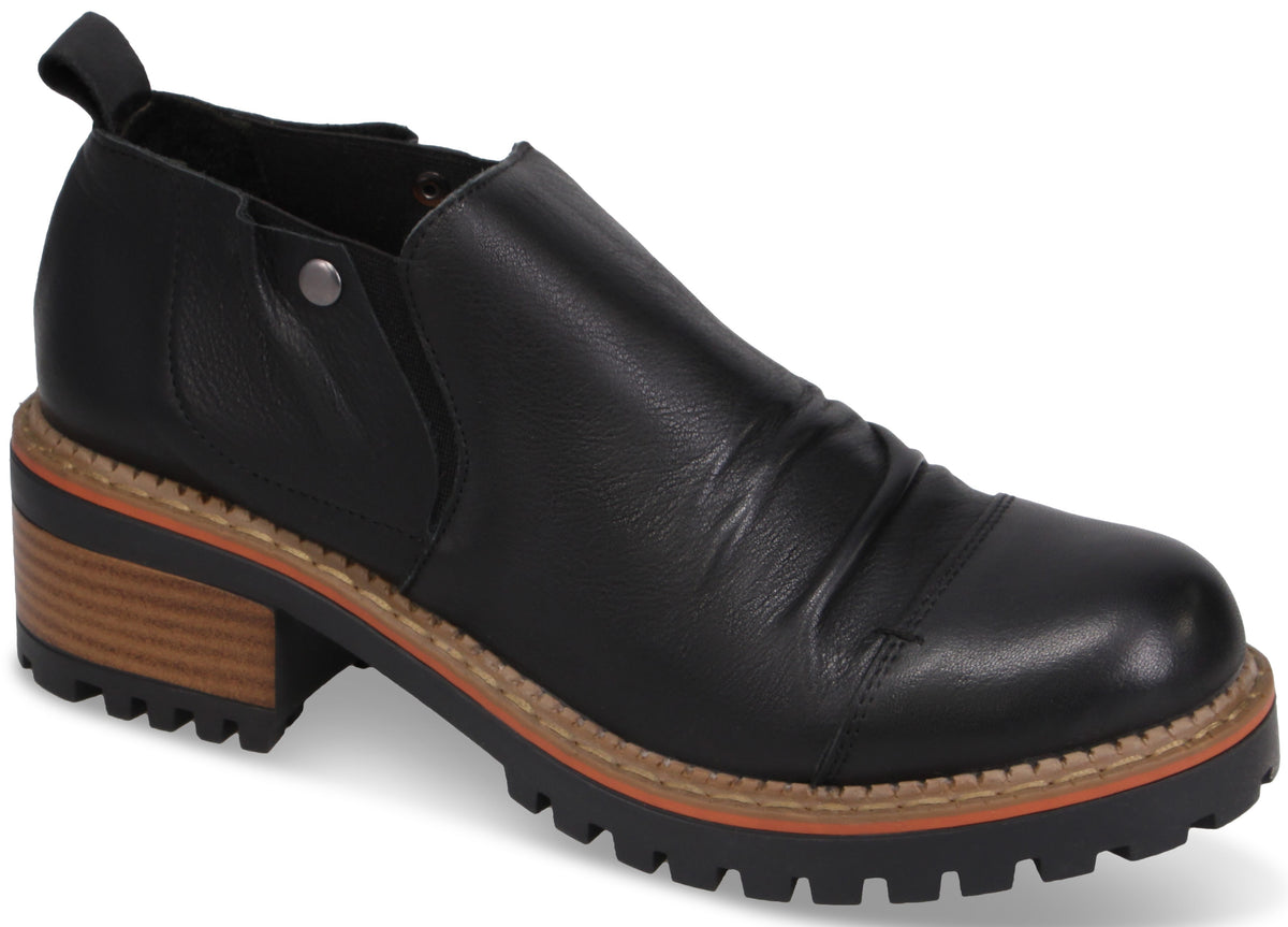A black slip-on ankle boot with a rounded toe, featuring a textured leather upper with subtle ruching, a silver decorative rivet on the side, a pull tab at the heel, and a thick, lug-style rubber outsole with a stacked wood-look heel and a thin orange welt stripe.