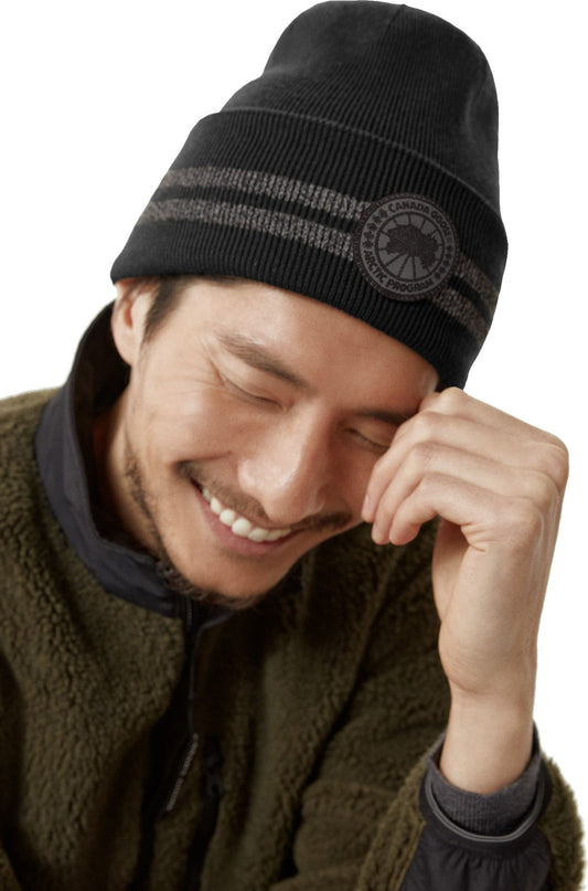 Person wearing a black knit beanie with two horizontal gray stripes and a circular logo.