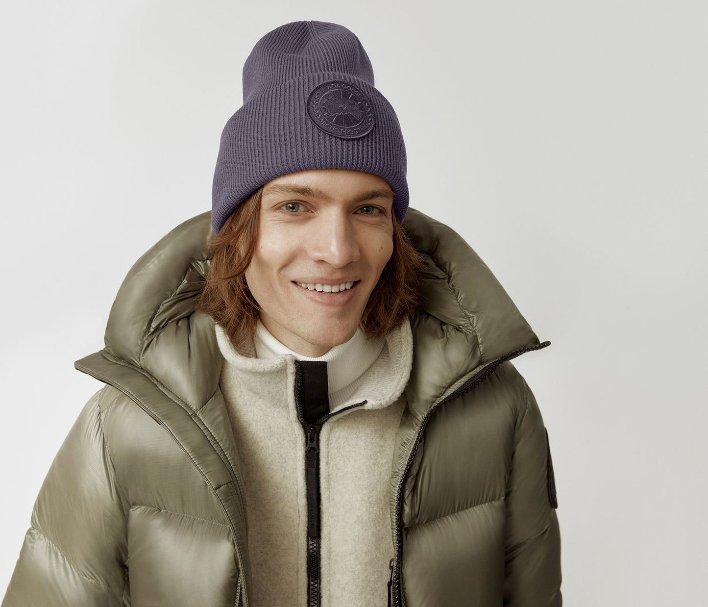 A person wearing a beanie, a light beige fleece, and a shiny green puffy jacket, smiling.