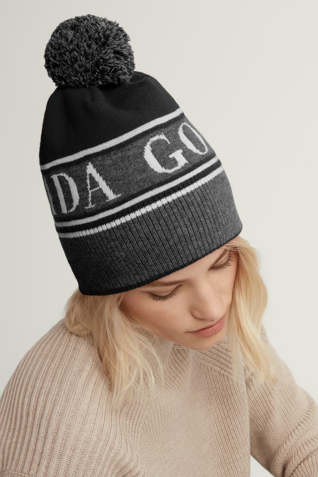 A gray and black beanie with white lettering and a gray pompom is on a fair-skinned woman with blonde hair.