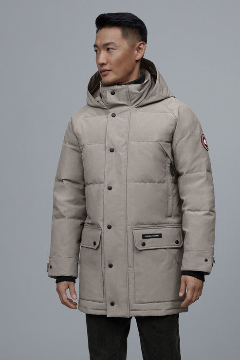 A man wearing a beige, mid-thigh length puffer jacket with a hood, front pockets, and button closure.