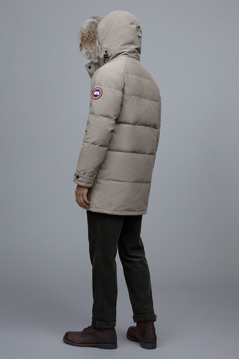 Person wearing a tan, mid-thigh length puffer coat with a hood trimmed with fur.