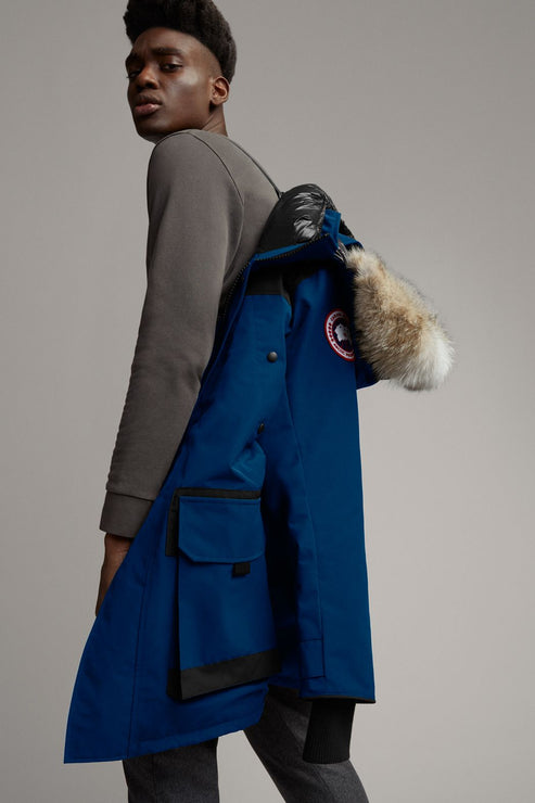 A man wearing a gray long sleeve shirt and gray pants is holding a blue parka over his shoulder, which has a fur-lined hood and front pockets.