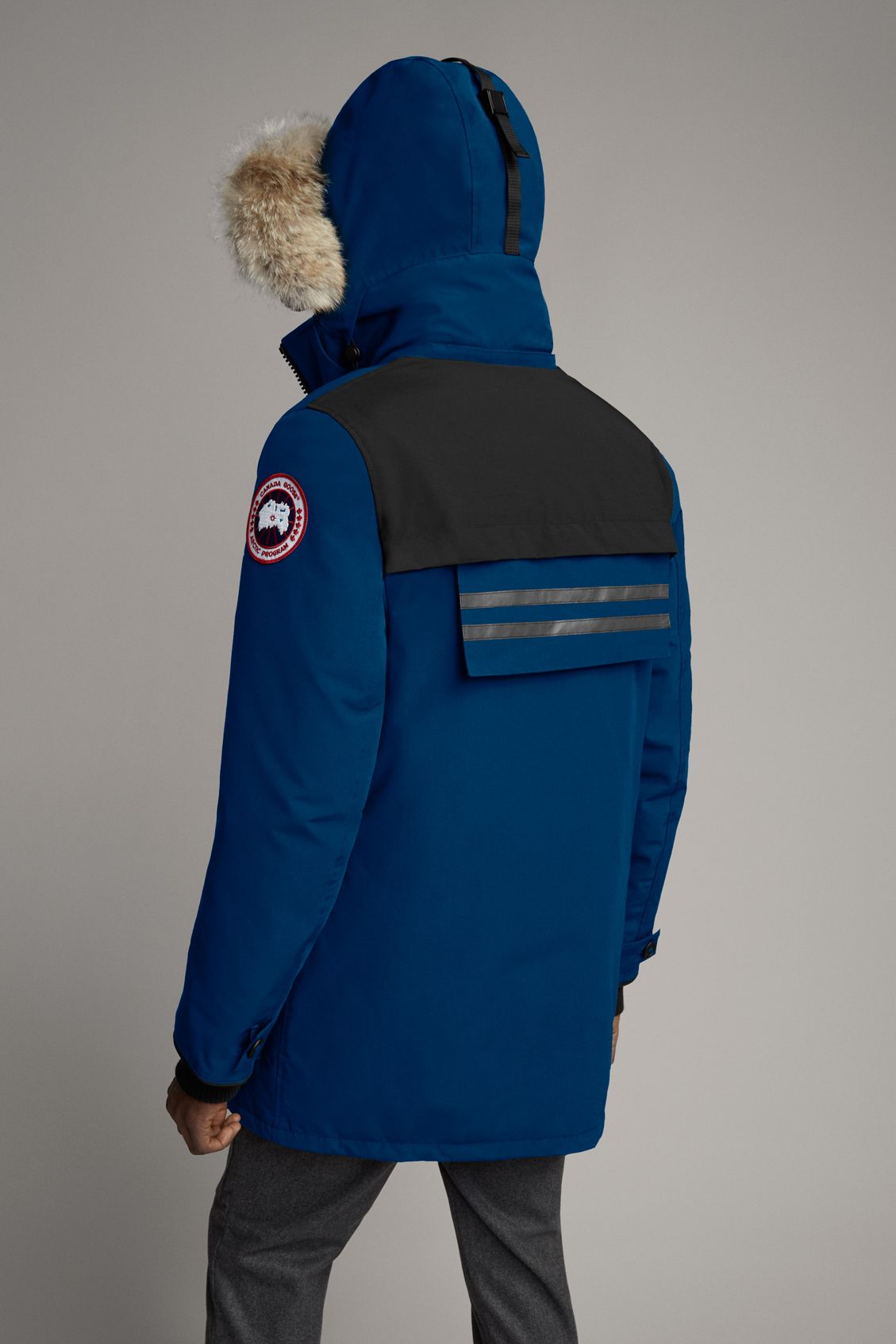 Rear view of a blue hooded parka with a fur ruff, a black yoke, reflective stripes, and a circular patch logo on the arm.