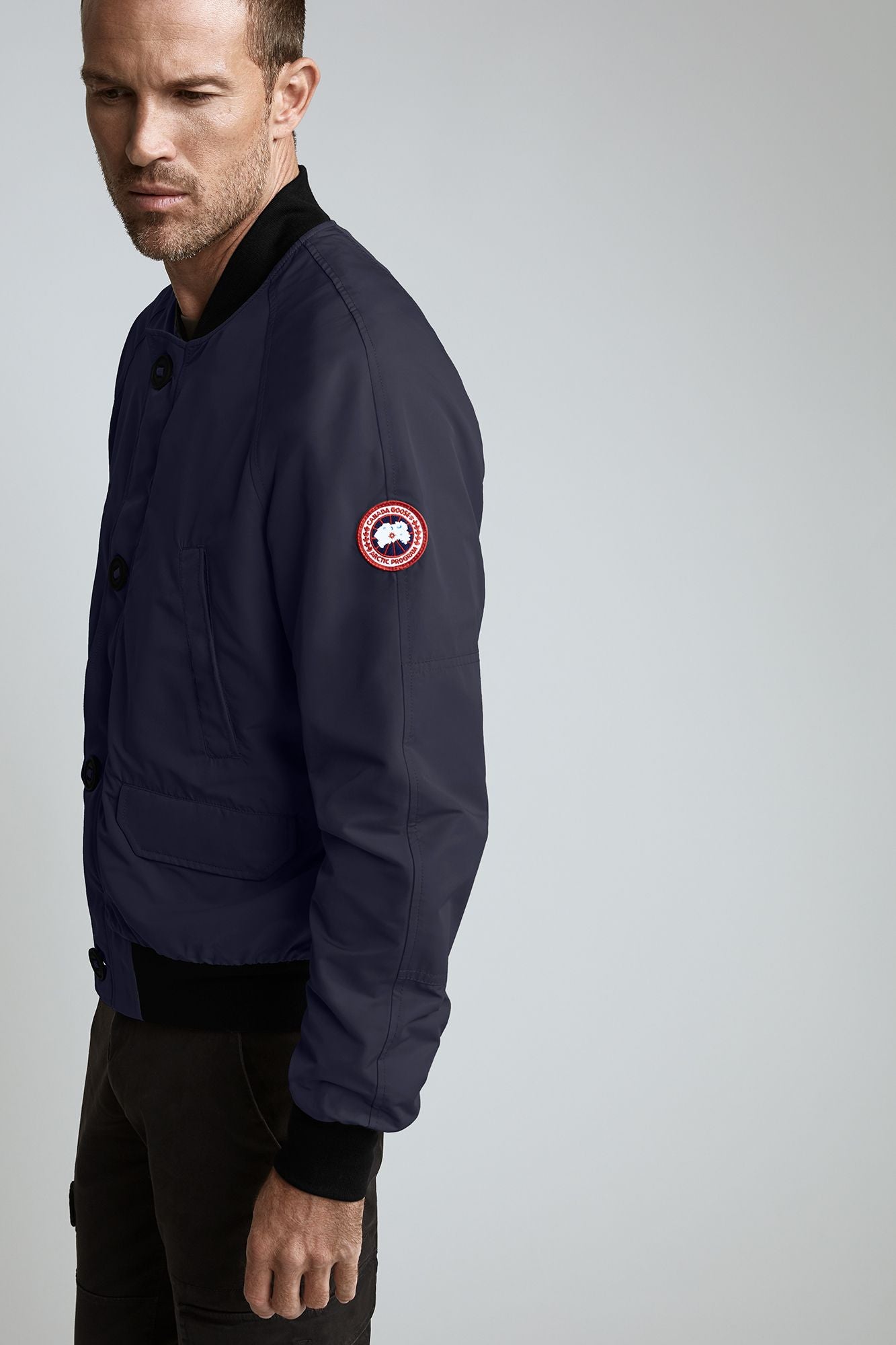 A man wearing a navy bomber jacket with a round logo on the upper left sleeve.