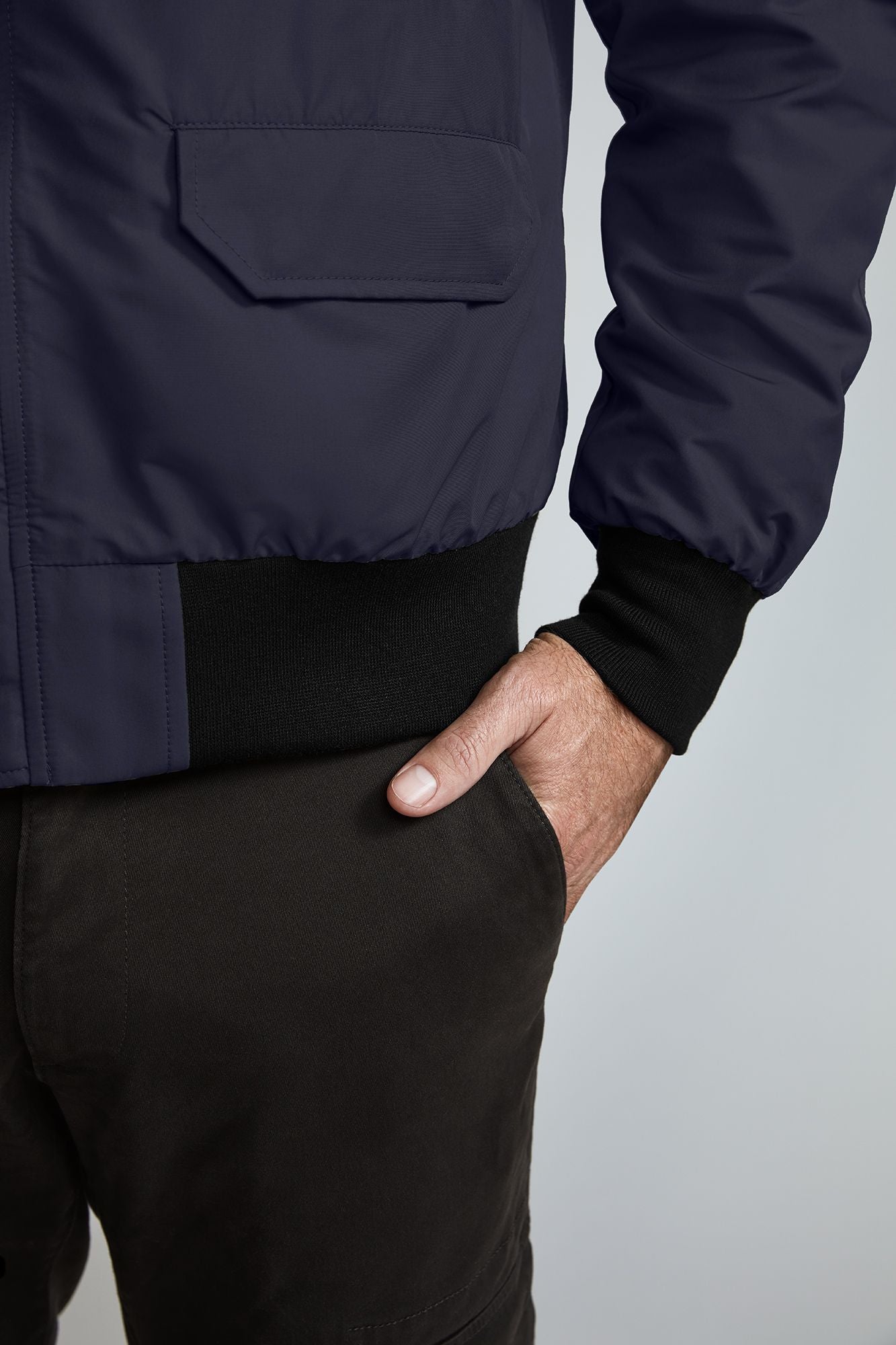 A close-up of a person's side with their hand in the pocket of their dark pants. The person is wearing a dark blue bomber-style jacket with black knitted cuffs and a chest pocket.