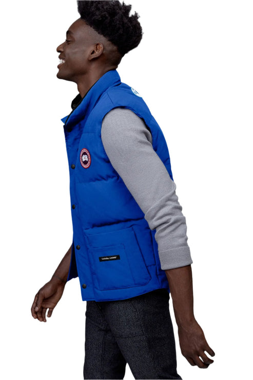 A man wears a blue, button-up puffer vest with the Goose logo on the upper left chest.